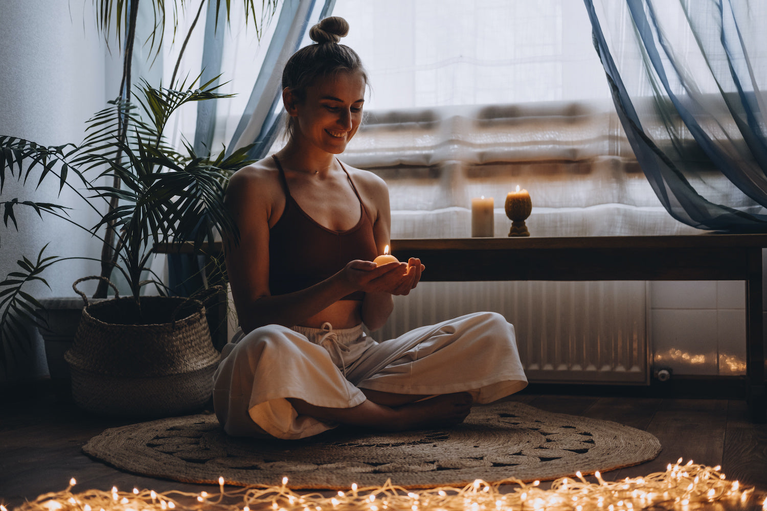 A woman in a tank top and linen pants, sitting cross, legged on a circular jute rug; smiling at a lit candle, sitting in front of a curtain window with a bench and lit candles in front of potted plants, with twinkle lights in the foreground.