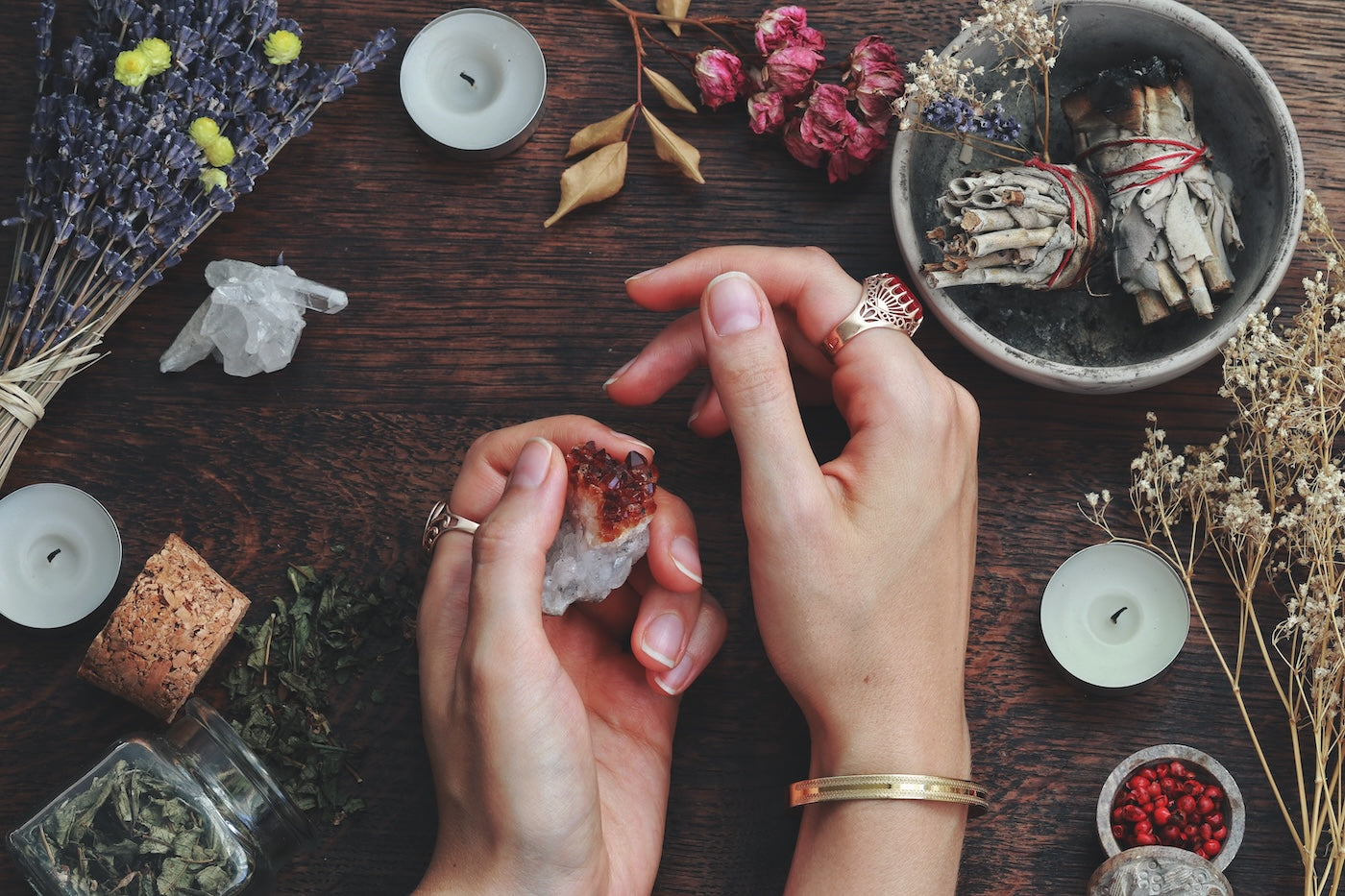 Lavender, candles, clear, quartz, cork, glass bottle with Herbs, dried flowers, smudging, sage stick, and hands with rings and bracelet, holding citrine crystal. A clearing, cleansing ritual tools.
