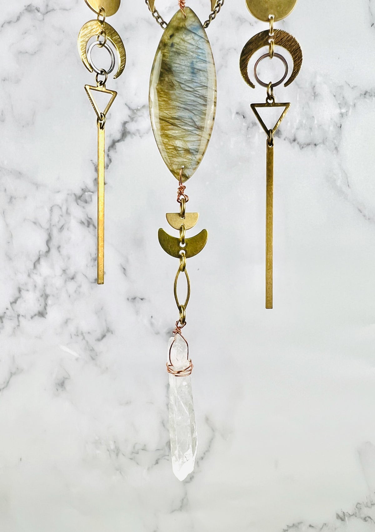 Three hanging vertical patterns depicting the bottom portion of a piece of crystal wall, art, including a gray labradorite, marquise shaped stone, and various brass shapes, including half circles, crescent, moons, a silver ring, triangles, and gold bars with a copper wire wrapped quartz crystal point hanging from the bottom. All hangs in front of a gray and white marble wall.