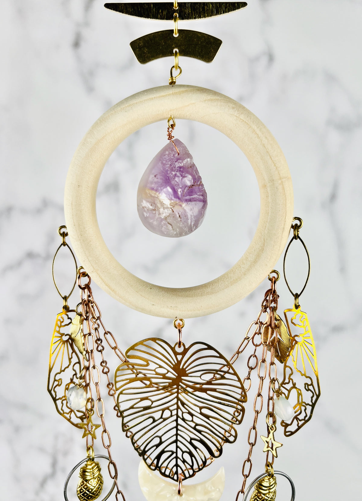 A hanging cream colored wood ring, connected to gold medal components at the top, and an amethyst teardrop shaped Bead in the center. From the bottom of the wood ring, hangs various metal shapes, including hand, outlines with sun and clouds in them, Opalite beads, copper colored chain, a large Monstera leaf, and acrylic cream, colored crescent moon, stars, silver rings, and pineapple charms; hanging in front of a gray and white marble wall.