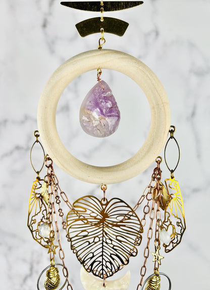 A hanging cream colored wood ring, connected to gold medal components at the top, and an amethyst teardrop shaped Bead in the center. From the bottom of the wood ring, hangs various metal shapes, including hand, outlines with sun and clouds in them, Opalite beads, copper colored chain, a large Monstera leaf, and acrylic cream, colored crescent moon, stars, silver rings, and pineapple charms; hanging in front of a gray and white marble wall.