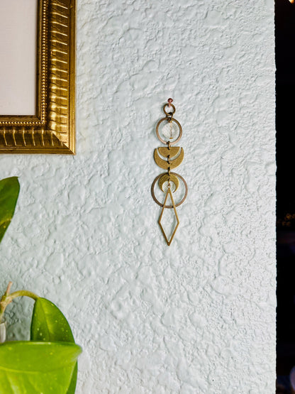 A piece of vertical clear quartz crystal art, connected by small rings with components such as a faceted Bead, bronze circles, gold colored half moons and crescent moons and a long diamond shape dangling from the bottom. The piece hangs on a light blue textured wall next to a gold frame and Plant.