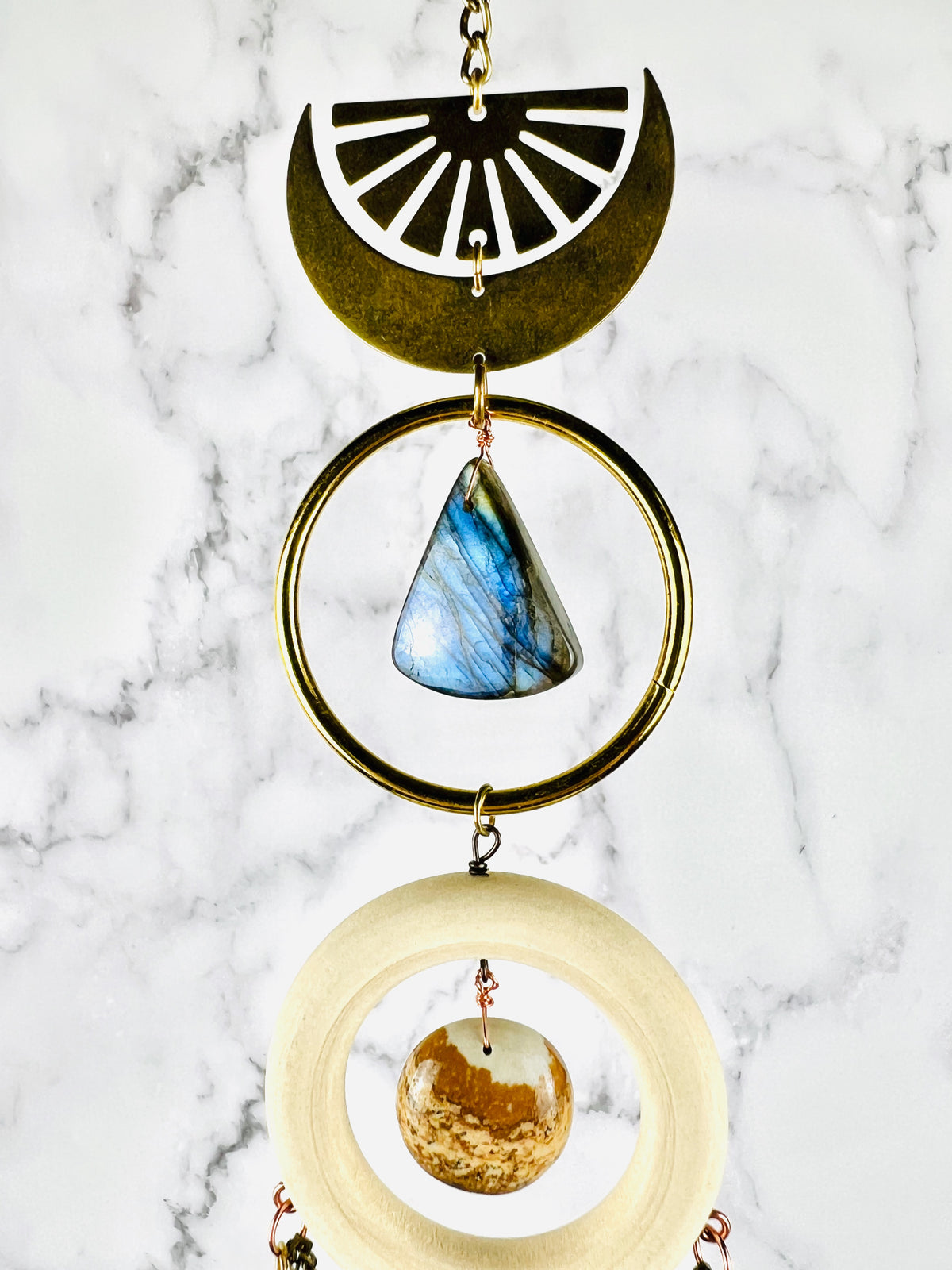 A beautiful iridescent, blue labradorite triangle shaped stone, suspended in a brass ring. Above it a crescent moon with a upside down sunburst and below it, a cream colored wood ring with a lovely piece of picture Jasper in light browns shades; all hangs in front of a marble background.