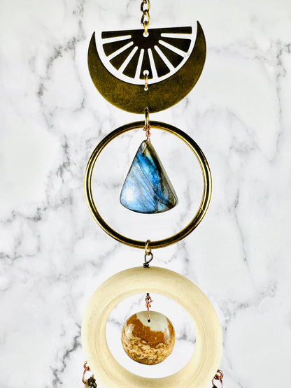 A beautiful iridescent, blue labradorite triangle shaped stone, suspended in a brass ring. Above it a crescent moon with a upside down sunburst and below it, a cream colored wood ring with a lovely piece of picture Jasper in light browns shades; all hangs in front of a marble background.