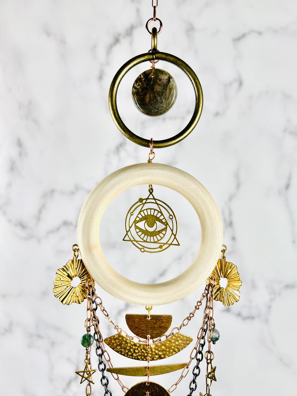 Vertically hanging crystal wall art featuring brass rings, labradorite crystals, a light wood ring with an eye charm surrounded by a triangle at its center. Hanging from this Charm, as copper colored chain, stars, moons and half circle gold colored metal shapes. The piece of art hangs in front of a marble wall.