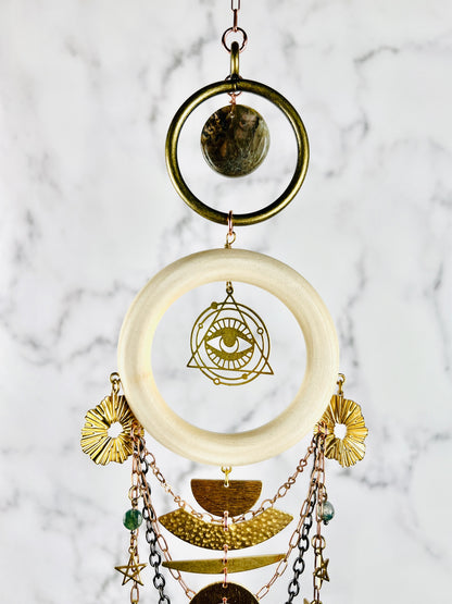 Vertically hanging crystal wall art featuring brass rings, labradorite crystals, a light wood ring with an eye charm surrounded by a triangle at its center. Hanging from this Charm, as copper colored chain, stars, moons and half circle gold colored metal shapes. The piece of art hangs in front of a marble wall.