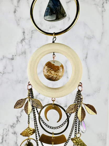 A beautiful piece of rounded triangle labradorite hangs in the middle of a brass ring, attached below it, a light wood ring with a center stone of picture Jasper in brown shades. Hanging below that, on the wood ring are bronze chains, and various metal shapes, such as leaves, fans, crescent, moons, and rings.