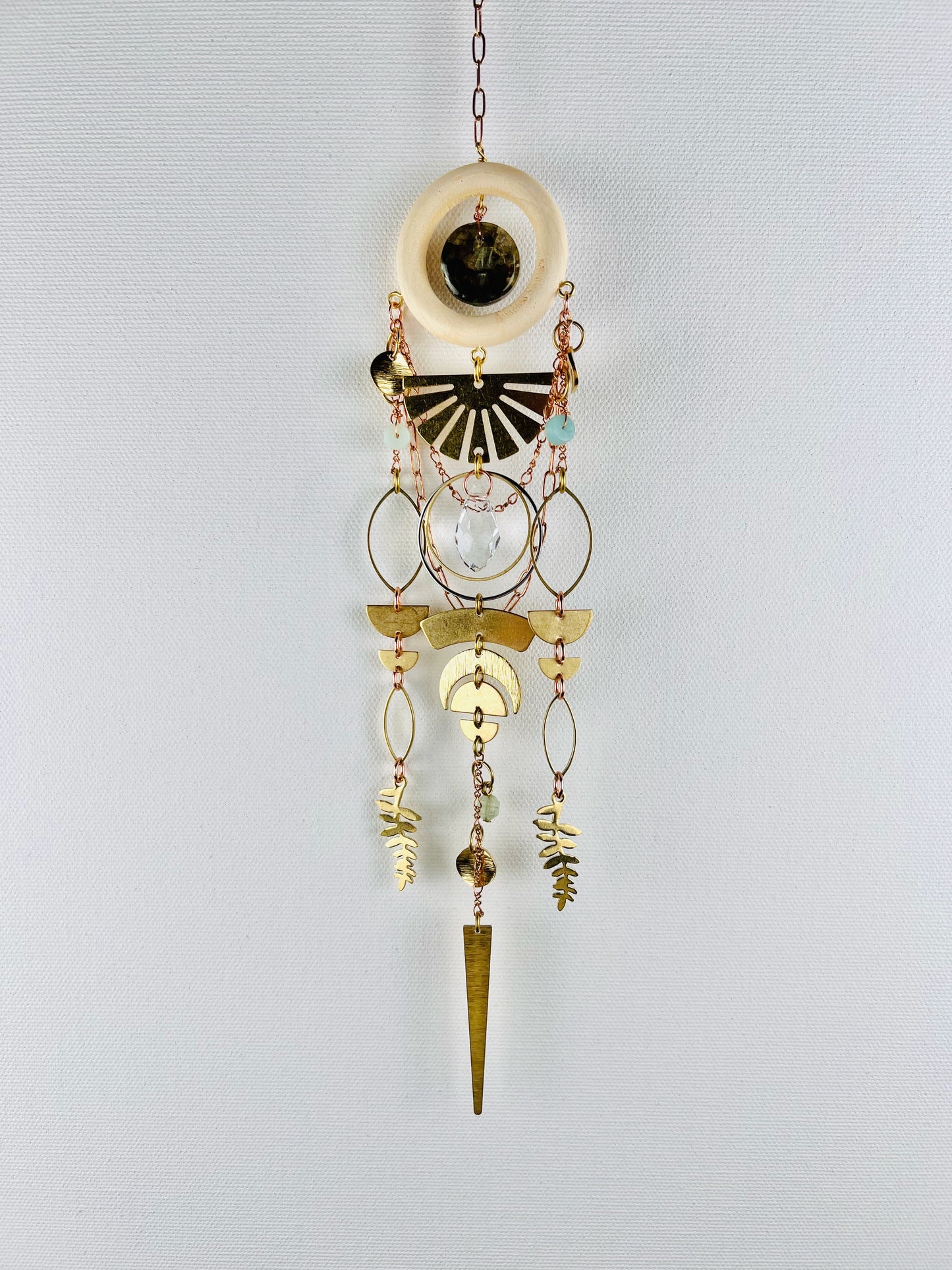 Decorative hanging crystal wall art with gold eye shapes, crescent moons, half circles, rings, leaves and amazonite, labradorite, prehnite and prism beads and geometric shapes on a light gray background