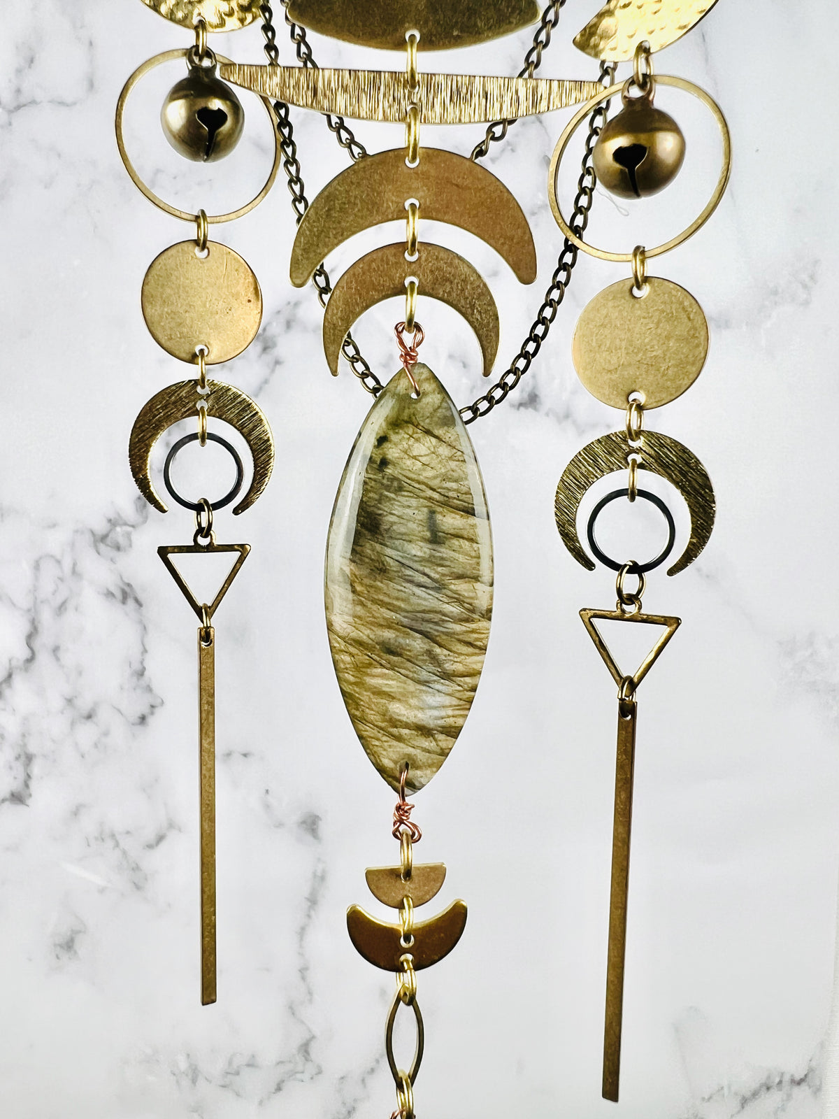 A lovely grouping of three hanging gold colored metal patterns linked by jump rings, including: brass bells, rings, circles, crescent moons, triangles, gold bars, and a marquee shaped labradorite crystal; all hanging in front of a gray and white marble background.