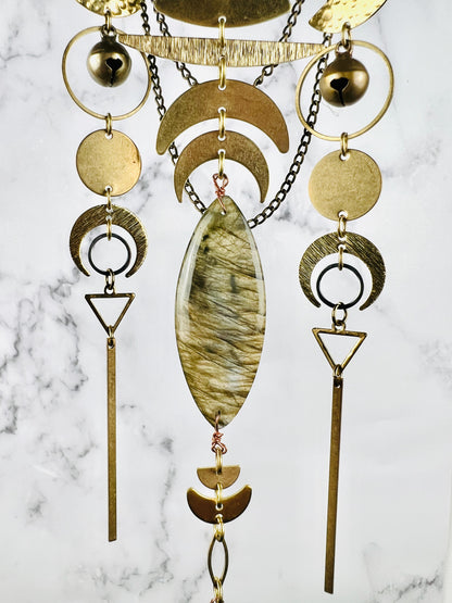 A lovely grouping of three hanging gold colored metal patterns linked by jump rings, including: brass bells, rings, circles, crescent moons, triangles, gold bars, and a marquee shaped labradorite crystal; all hanging in front of a gray and white marble background.