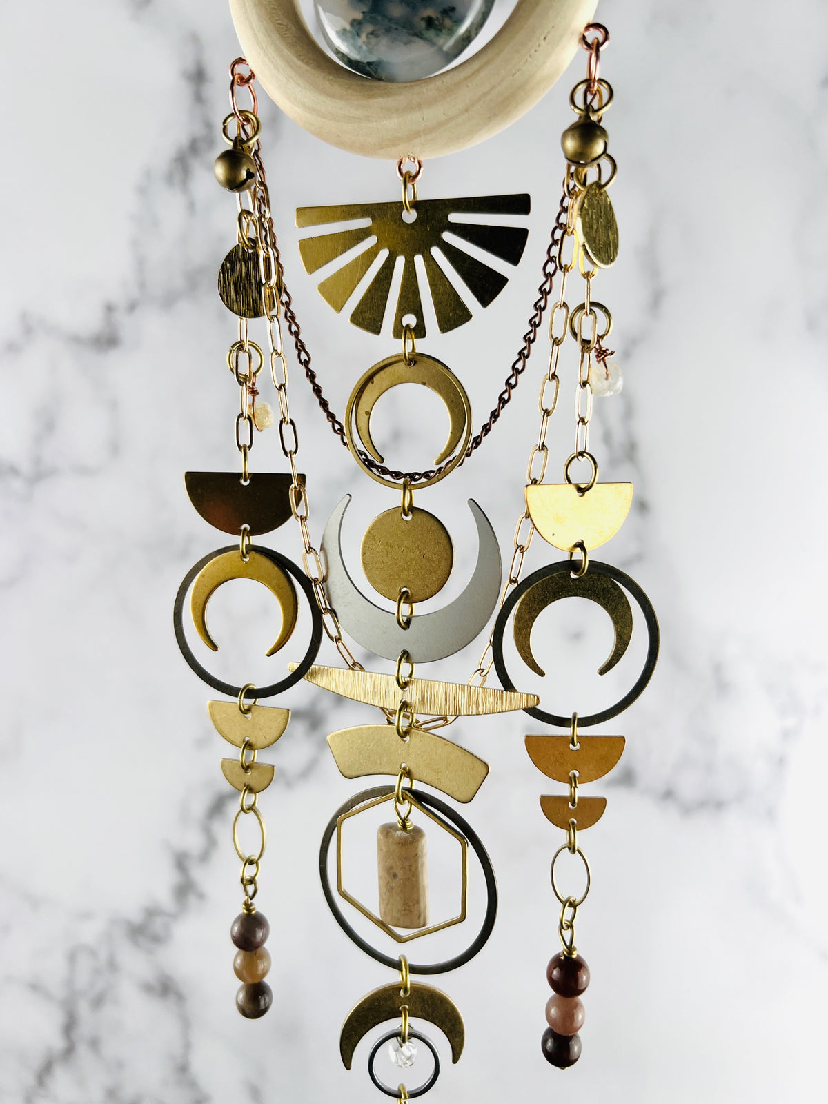 middle view of hanging crystal wall art made of wood, moss agate, half circles, crescent, moons, circles, rings, oval, hexagons, chain and wire wrapped, Herkimer, diamond, topaz, petrified Wood, Jasper and Moss agate beads. The piece hangs vertically in front of a marble wall.