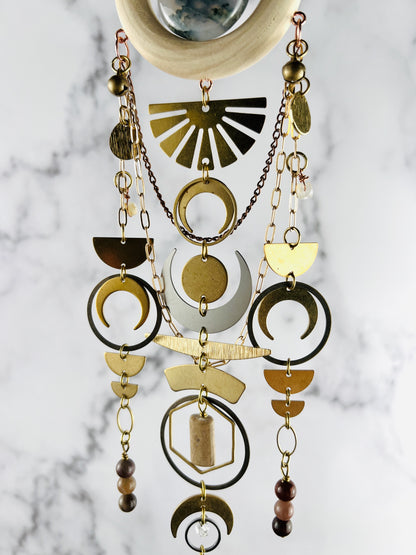 middle view of hanging crystal wall art made of wood, moss agate, half circles, crescent, moons, circles, rings, oval, hexagons, chain and wire wrapped, Herkimer, diamond, topaz, petrified Wood, Jasper and Moss agate beads. The piece hangs vertically in front of a marble wall.