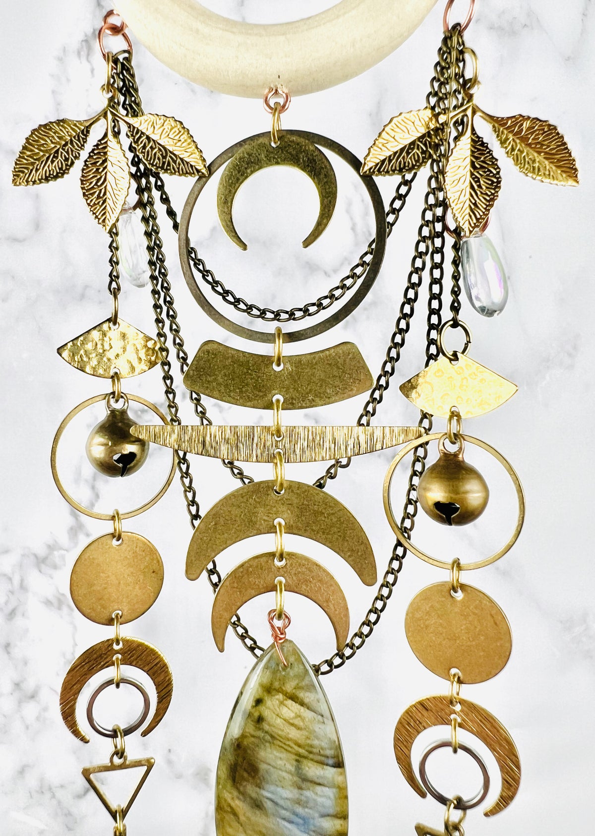 A beautiful arrangement of gray labradorite crystal and metal shapes including: crescent moons, leaves, bells, rings, circles, triangles, and fan shapes with a clear drop crystal attached on either side. I'll hang in front of a gray and white marble background.