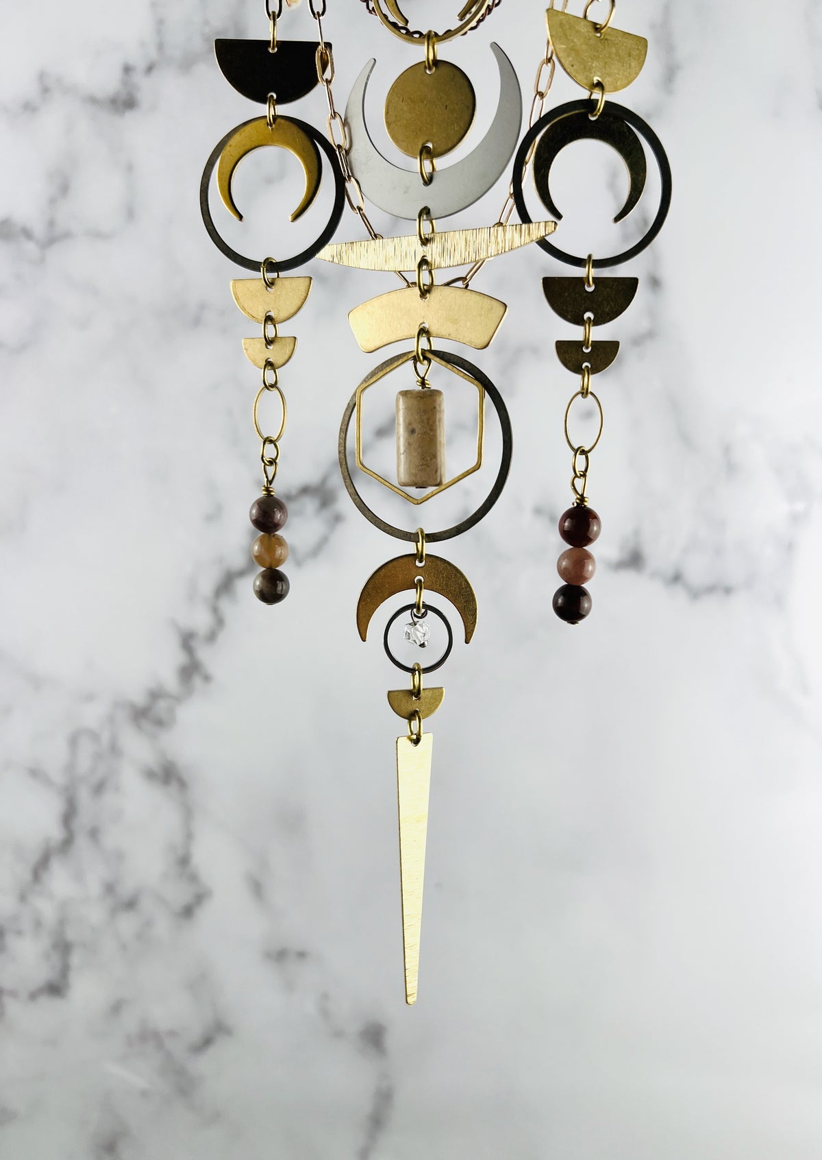 the bottom portion of a verticle art piece described a crystal wall art consisting of three connected patterns of a combination of metal charms including: half circles, crescent, moons, Rings, ovals, Jasper, petrified wood, Herkimer, diamond, beads, and triangles hanging in front of a marble wall.