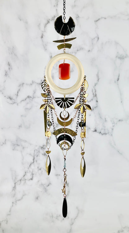 A vertically hanging piece of art with various materials, including: a cream colored wood ring, black obsidian, orange carnelian, blue aquamarine, and black Tourmaline crystals with gold colored mteal crescent moons, a sun burst, leaves, half circles, triangles, ovals, and stars adorn this Dreamcatcher like peace as it hangs in front of a gray and white marble wall.