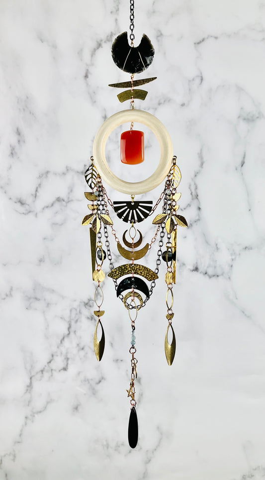 A vertically hanging piece of art with various materials, including: a cream colored wood ring, black obsidian, orange carnelian, blue aquamarine, and black Tourmaline crystals with gold colored mteal crescent moons, a sun burst, leaves, half circles, triangles, ovals, and stars adorn this Dreamcatcher like peace as it hangs in front of a gray and white marble wall.