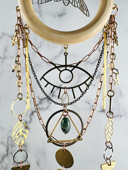 A decorative crystal wall hanging featuring a light wood ring at the top, and silver bug charm in the middle, suspended with copper and brass toned chains. It includes gold colored metal charms shaped like an eye, a triangle, stars, leaves, and circles, rings with a single green marbled stone bead on a marble background.