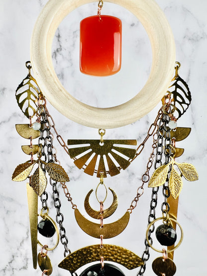 Upper section of a piece of carnelian crystal wall art that resembles a Dreamcatcher, including components, such as a cream colored wood ring, aquamarine and black tourmaline wire wrapped beads with various metal charms, such as leaves, half circles, and upside down sunburst, crescent moons, circle and rings with copper colored and gun metal colored chain attached to the bottom portion of the wood ring. This piece hangs in front of a gray and white marble wall.
