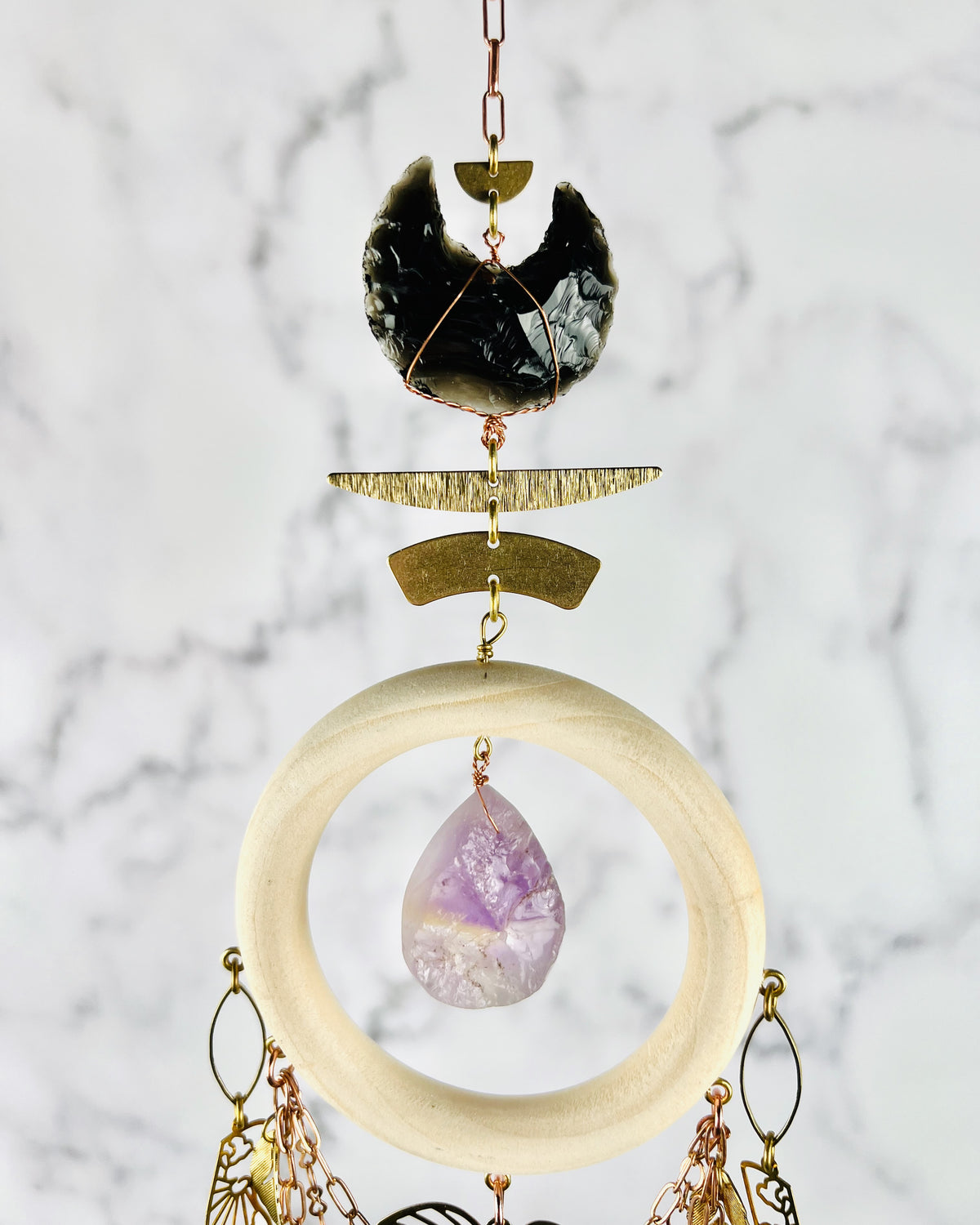 The top of a piece of vertically hanging crystal wall, art, including copper, colored chain, half circle, crescent, moon, obsidian, wire, wrapped crystal, brass metal pieces, a light wood ring with a amethyst, hanging in the middle and chain and metal charms hanging from the wood. It all hangs in front of of a marble wall.