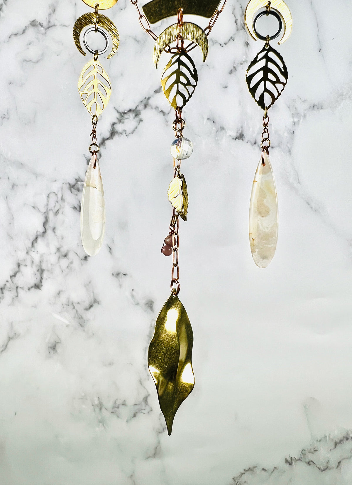 From the top of the photo hangs the bottom portion of a piece of crystal wall, art, including: gold colored metal, crescent moons, silver rings, gold metal leaves, wire wrapped flower agate drop shaped crystals, a faceted quartz, and lepidolite beads. At the very bottom of this art hangs a bending golden leaf, and the entire Charm hangs in front of a gray and white marble wall.