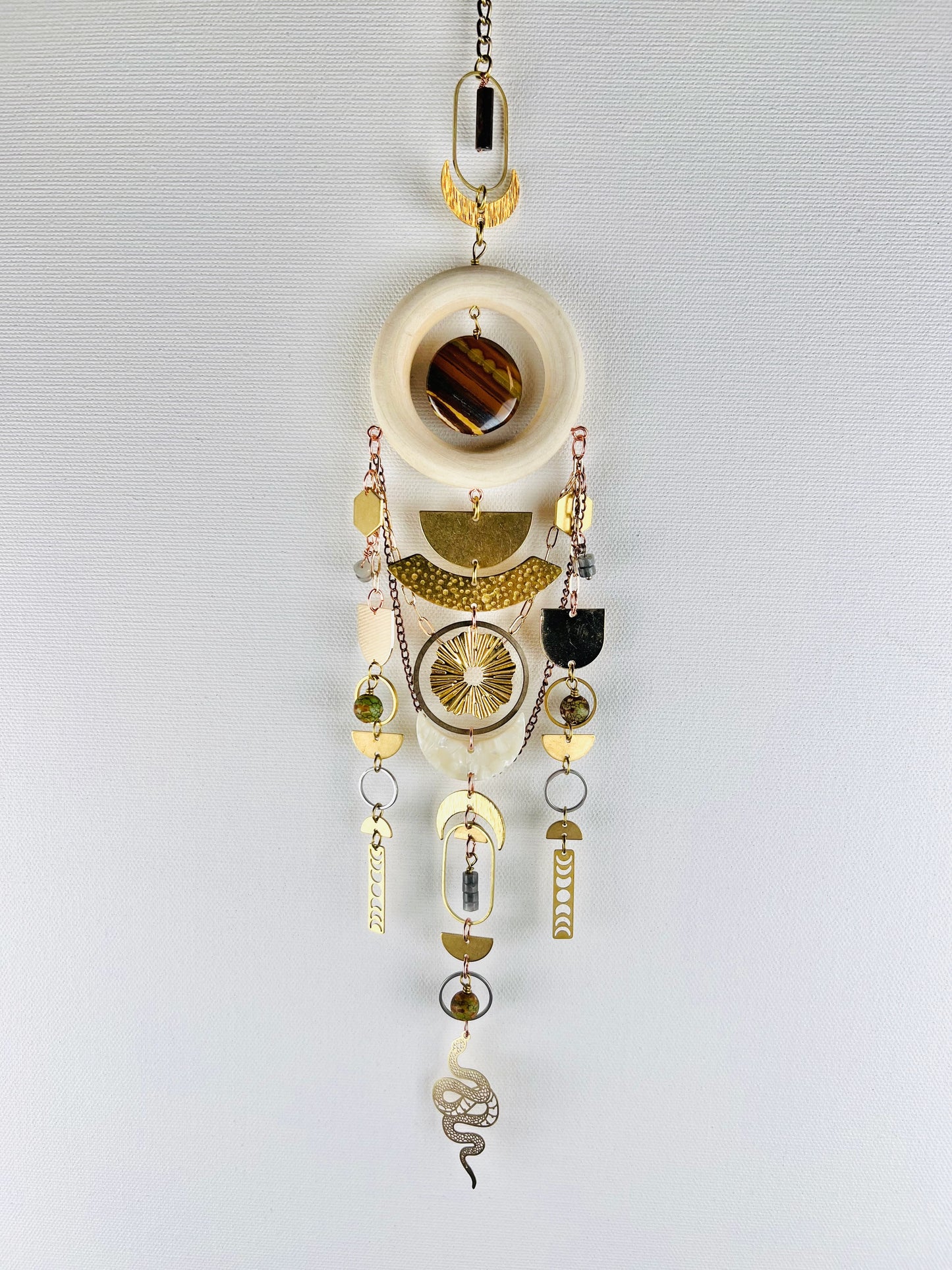 Decorative hanging crystal wall art with gold half circles, rings, ovals, moon phase, sun burst, crescent moons, hexagons, chain, silver rings, copper rings and wood ring, also snake and beads on a white background