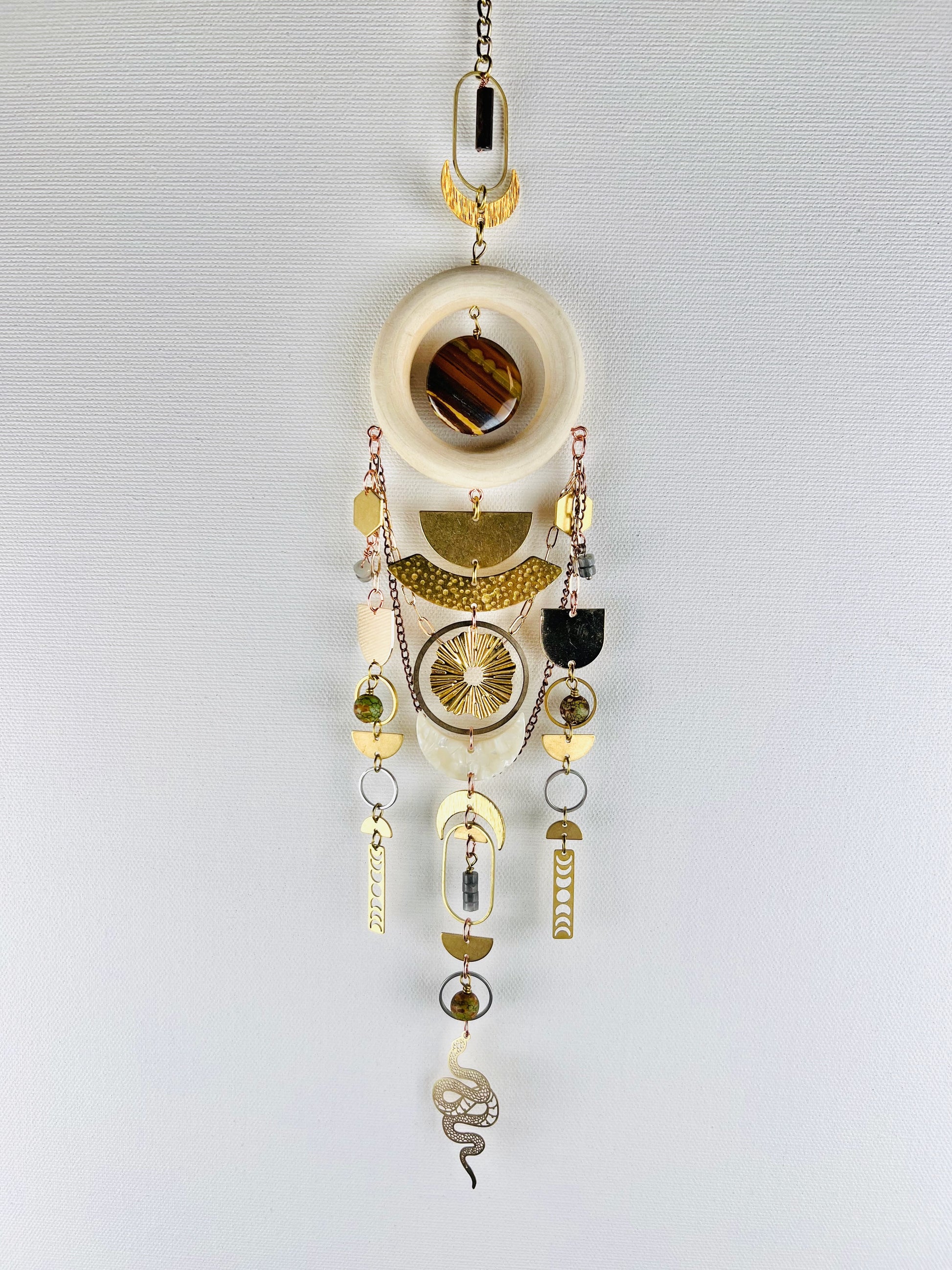 Decorative hanging crystal wall art with gold half circles, rings, ovals, moon phase, sun burst, crescent moons, hexagons, chain, silver rings, copper rings and wood ring, also snake and beads on a white background