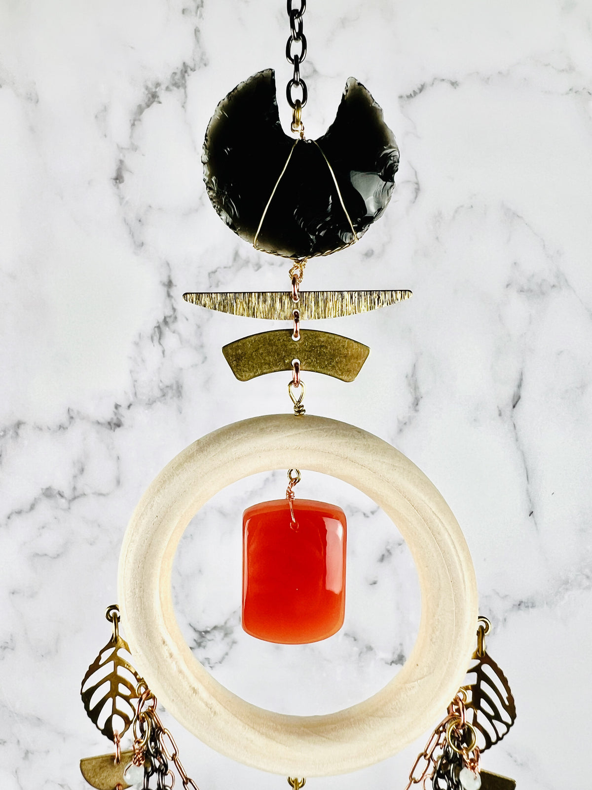 A Dreamcatcher like piece of hanging crystal wall art with deep meaning and a crystal focused theme. This piece hangs from gold colored chain, followed by an obsidian crescent moon crystal with metal components below that and a cream colored wood ring attached to those with a center rounded pillow shaped carnelian stone. Attached to the wood ring are metal leaf shapes and copper colored chain as well as white wire wrapped beads.