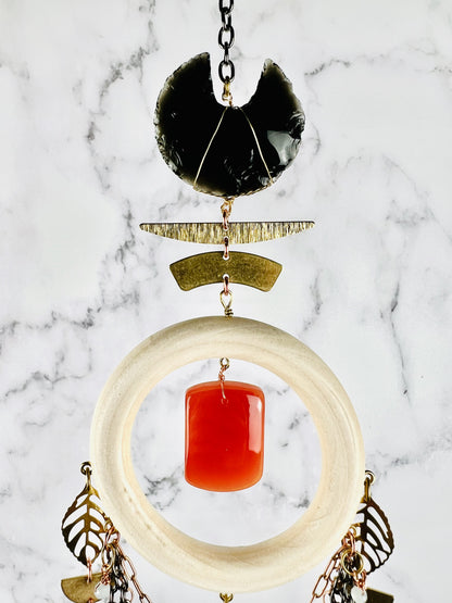 A Dreamcatcher like piece of hanging crystal wall art with deep meaning and a crystal focused theme. This piece hangs from gold colored chain, followed by an obsidian crescent moon crystal with metal components below that and a cream colored wood ring attached to those with a center rounded pillow shaped carnelian stone. Attached to the wood ring are metal leaf shapes and copper colored chain as well as white wire wrapped beads.