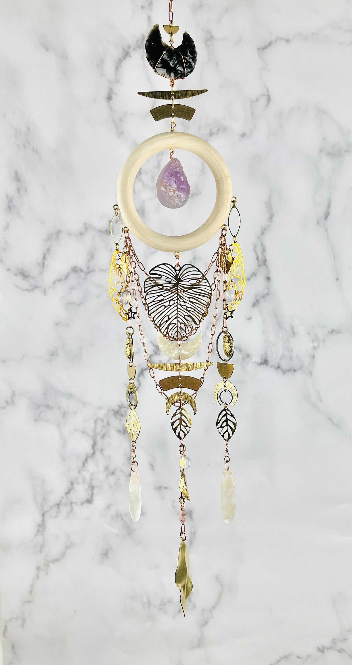 A bohemian dreamcatcher-style piece of wall art, featuring obsidian, purple amethyst, flower agate, Opalite, and lepidolite beads, hanging against a white and gray marble background. The art also features a cream-colored wooden ring, gold-toned metal elements, a large monstera leaf, and various dangling gold colored metal charms including leaves, moons, and beads.