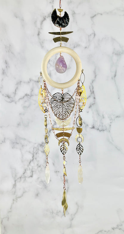 A bohemian dreamcatcher-style piece of wall art, featuring obsidian, purple amethyst, flower agate, Opalite, and lepidolite beads, hanging against a white and gray marble background. The art also features a cream-colored wooden ring, gold-toned metal elements, a large monstera leaf, and various dangling gold colored metal charms including leaves, moons, and beads.
