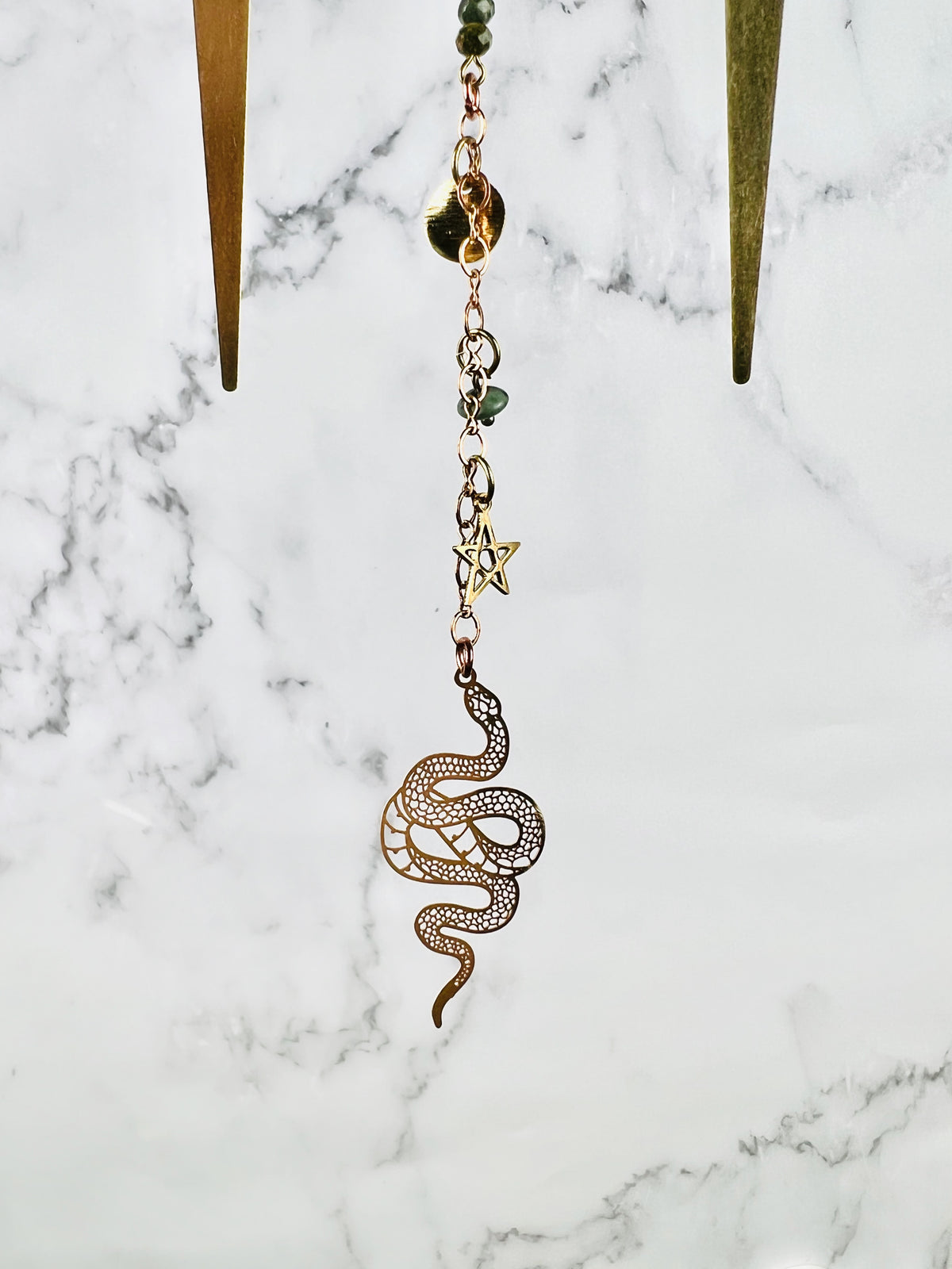 The bottom portion of a piece of crystal wall, art, including metal spikes, copper colored chain, wire wrapped beads, a star, and a snake charm all hanging in front of a marble wall.
