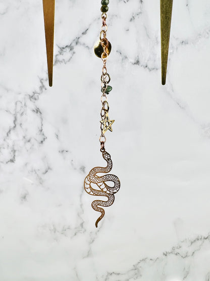 The bottom portion of a piece of crystal wall, art, including metal spikes, copper colored chain, wire wrapped beads, a star, and a snake charm all hanging in front of a marble wall.