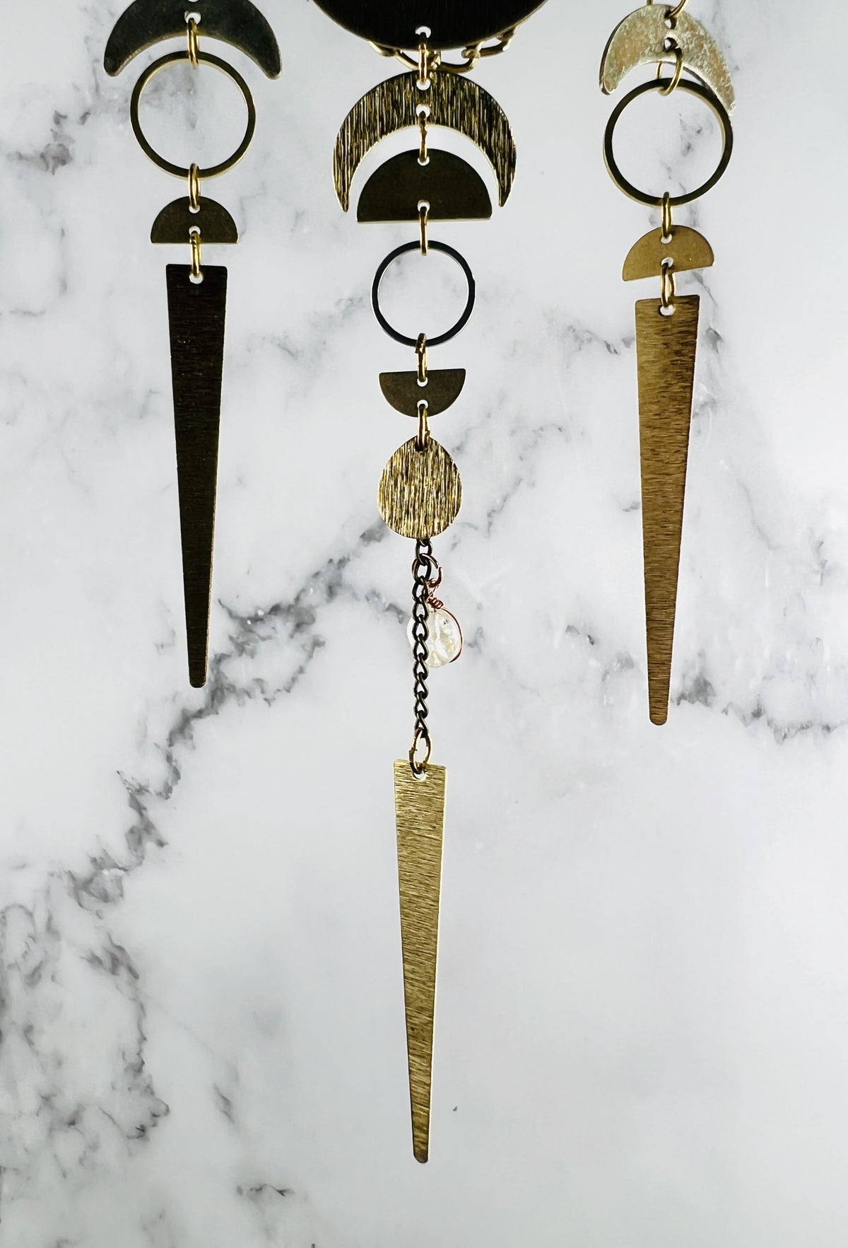 vertically hanging wall art segments with metal charms including crescent moons, half circles, rings, chain and topaz hanging in front of a marble background. 