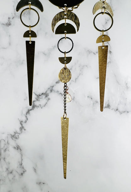 vertically hanging wall art segments with metal charms including crescent moons, half circles, rings, chain and topaz hanging in front of a marble background. 
