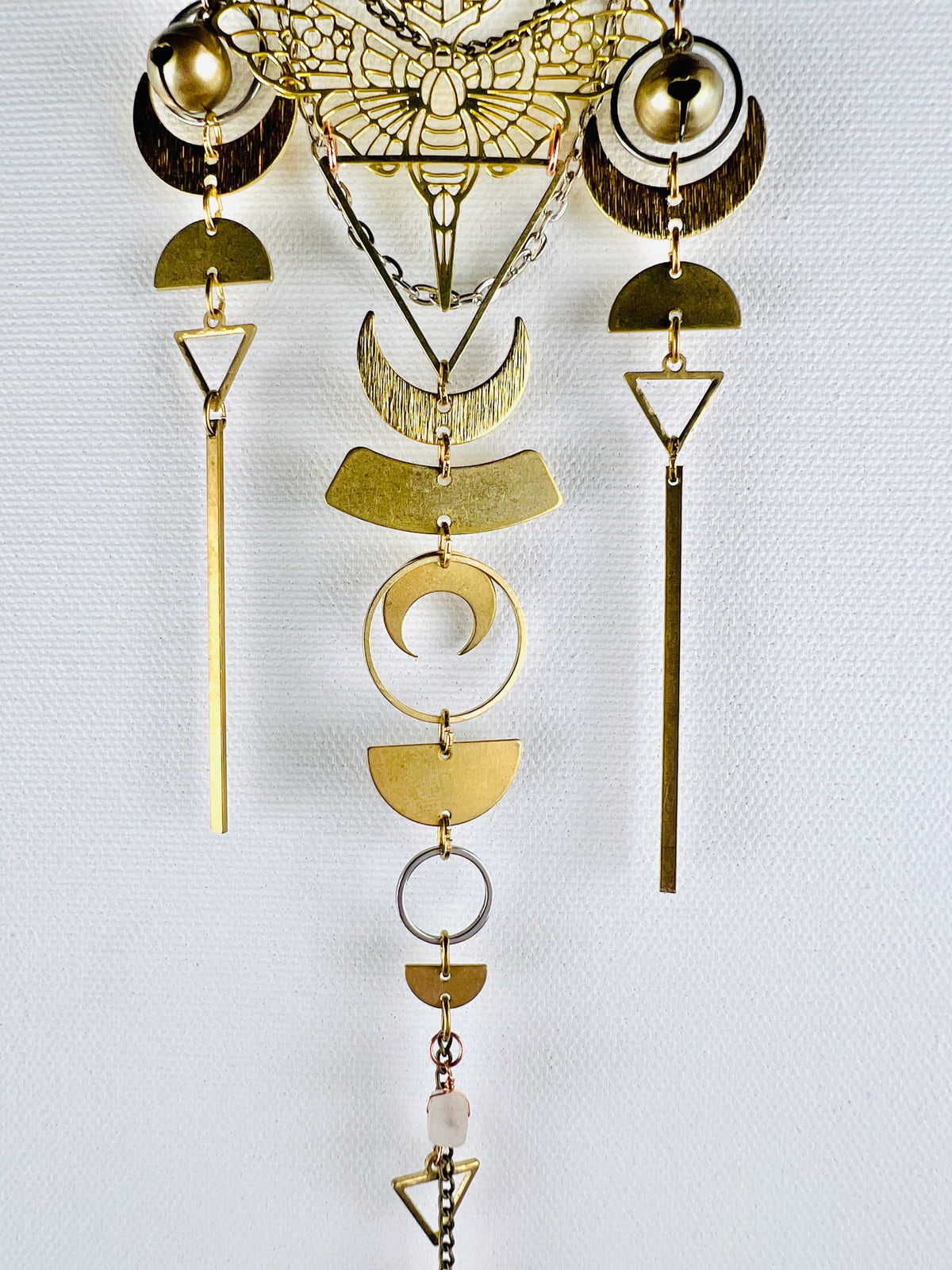 Three vertical lines of beautifully designed, gold-colored metal shapes hang in this photo, including: a moth, brass bells, silver rings, crescent, moons, half circles, triangles, a wire wrapped piece of rose, quartz, and long gold bars. They are all attached by tiny gold Rings and hang in front of and light gray wall.