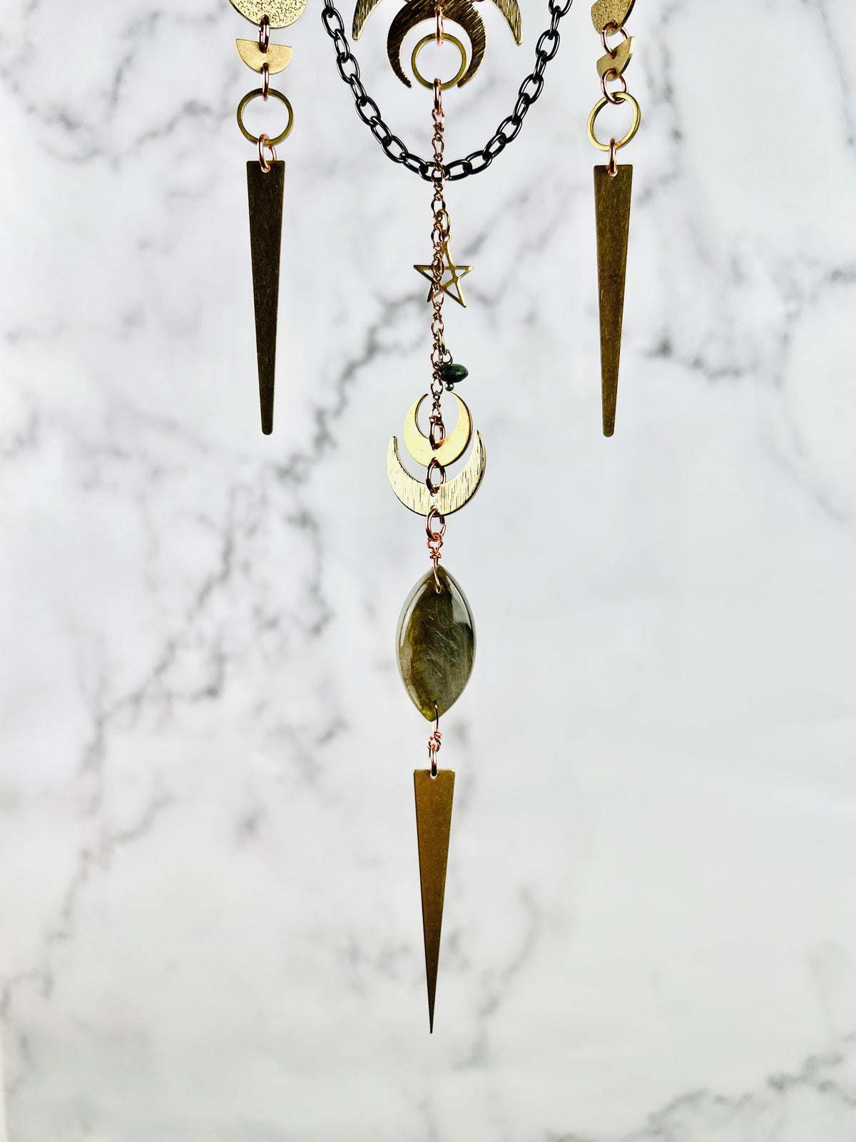 The bottom portion of a hanging crystal wall, art piece with crescent moons, half circles, rings, long triangles, star, chain, and a labradorite eye shaped bead, hanging in front of a marble wall.