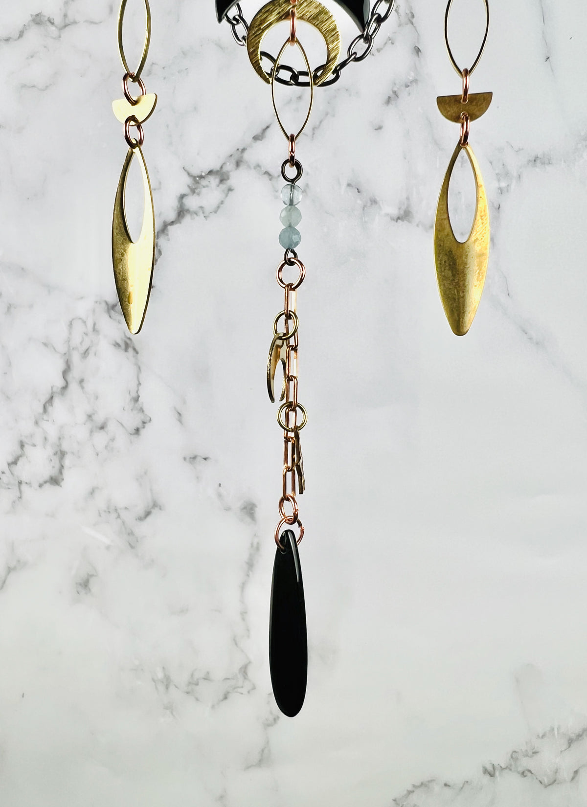 A close-up photo of three separate hanging metal component designs,attached by Rings And chain. Obsidian and aquamarine beads adore this piece along with gold colored metal, ovals, half circles and a crescent moon Charm, the peace hangs in front of a gray and white marble wall.