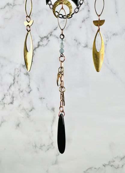 A close-up photo of three separate hanging metal component designs,attached by Rings And chain. Obsidian and aquamarine beads adore this piece along with gold colored metal, ovals, half circles and a crescent moon Charm, the peace hangs in front of a gray and white marble wall.