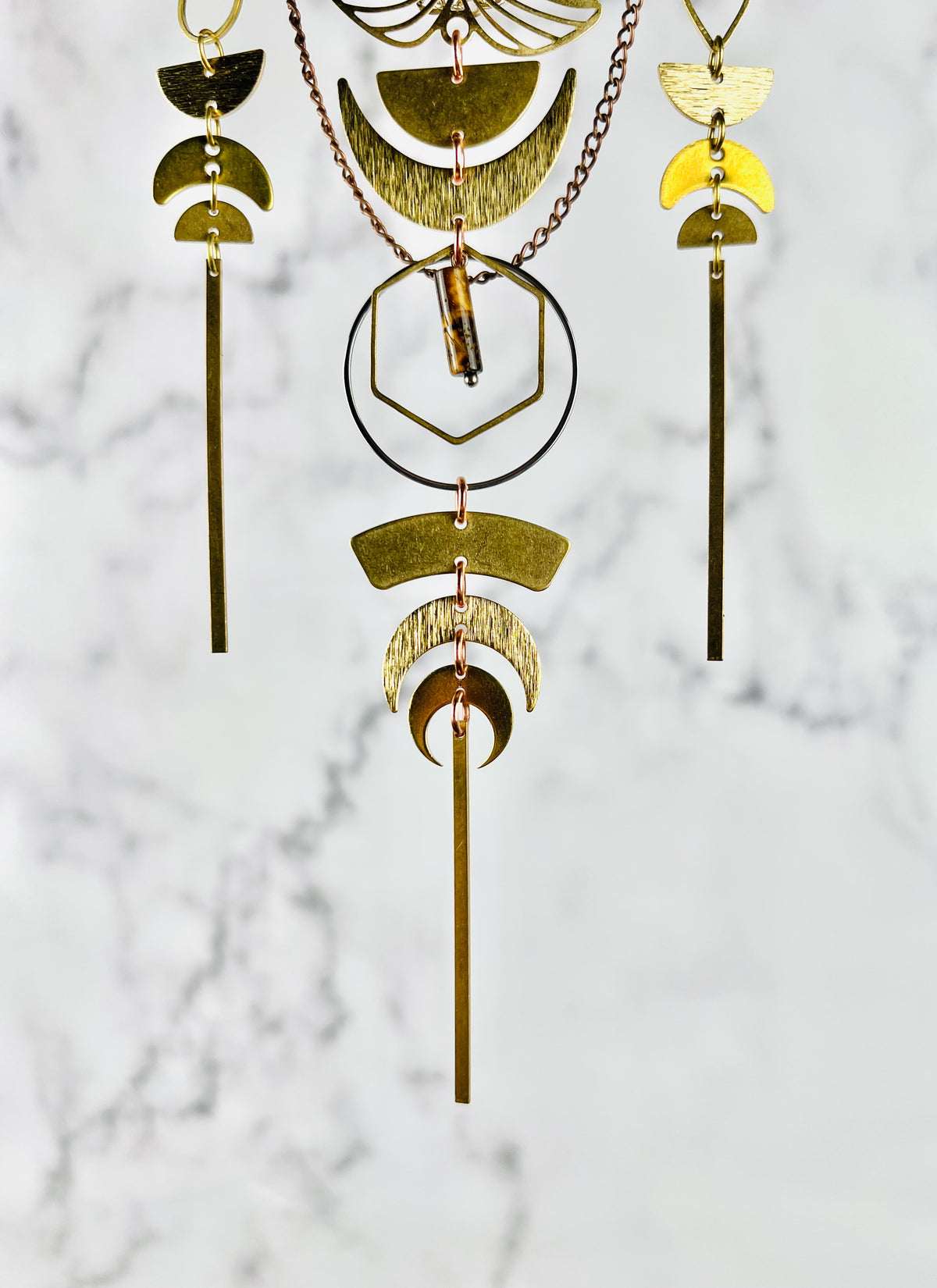 Three groupings of hanging, connected metal Moon phases, with chain and geometric shapes including half circles, crescent, moons, long bars, hexagons, and rings, in gold and silver metal tones hanging in front of a marble background.