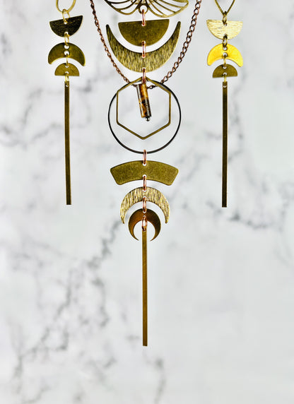 Three groupings of hanging, connected metal Moon phases, with chain and geometric shapes including half circles, crescent, moons, long bars, hexagons, and rings, in gold and silver metal tones hanging in front of a marble background.