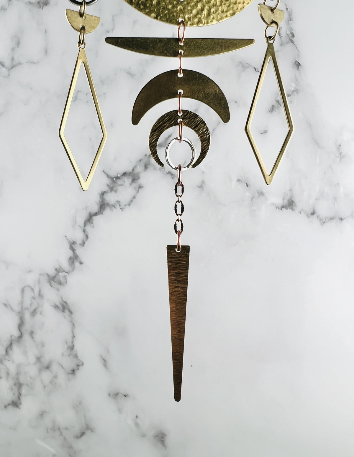 Two diamond shaped gold colored metal charms with crescent moons, copper colored chain, silver ring, and long metal triangle at the bottom hang in front of a marble wall. it is the bottom portion of an intricate piece of vertically hanging crystal wall art.