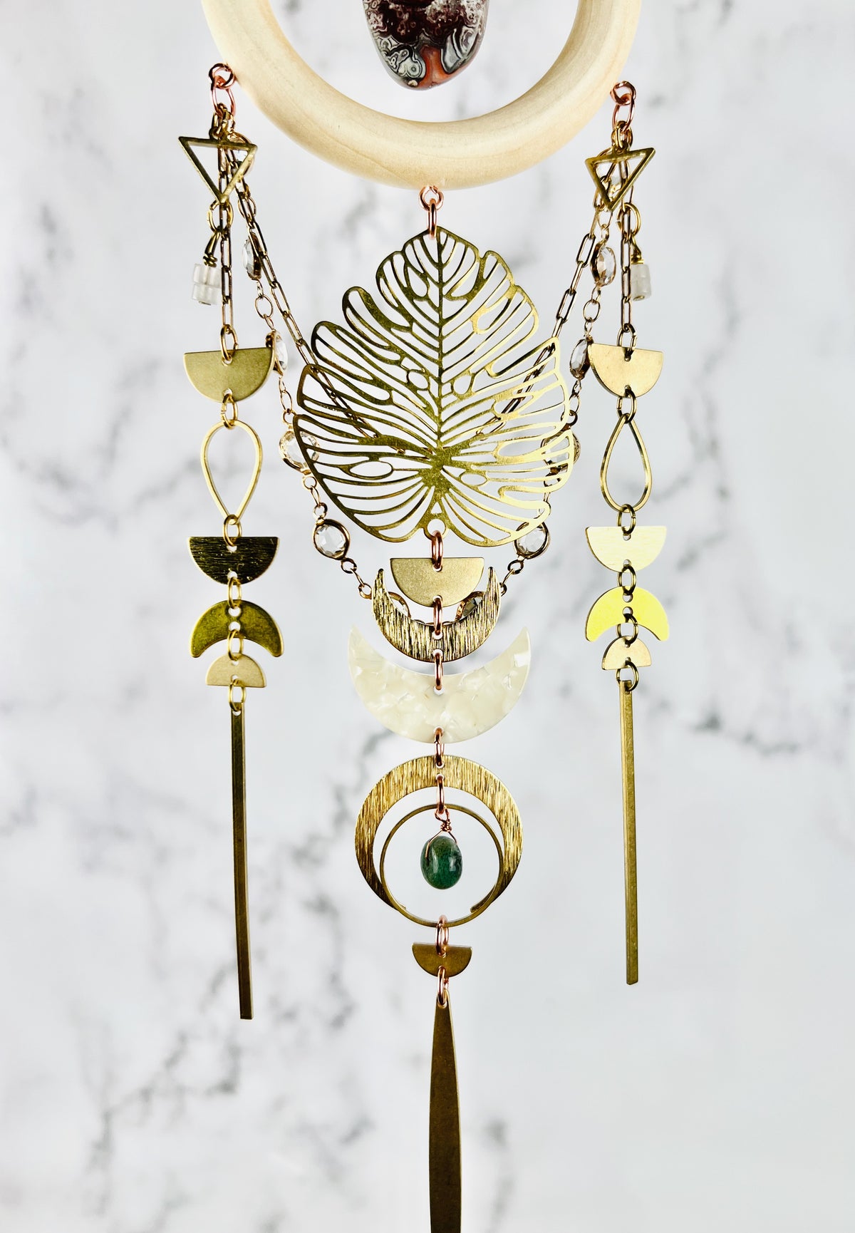 Vertically Hanging crystal wall art including, a cream colored wood ring with a red and gray, crazy lace agate stone in the middle of it. Attached to the bottom of the wood ring our metal components, such as: triangles, wire, wrapped beads, gold colored chain with prisms attached, half circles, teardrop shapes, crescent, moons, rings and long gold metal bars as well as a green moss agate Bead. This art piece hangs in front of a gray and white marble wall.