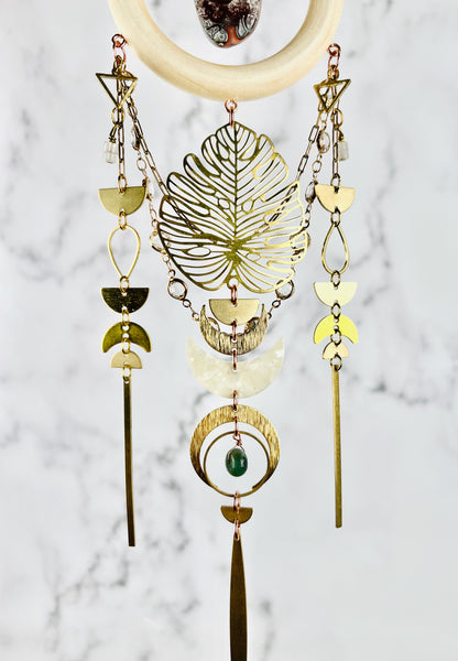Vertically Hanging crystal wall art including, a cream colored wood ring with a red and gray, crazy lace agate stone in the middle of it. Attached to the bottom of the wood ring our metal components, such as: triangles, wire, wrapped beads, gold colored chain with prisms attached, half circles, teardrop shapes, crescent, moons, rings and long gold metal bars as well as a green moss agate Bead. This art piece hangs in front of a gray and white marble wall.