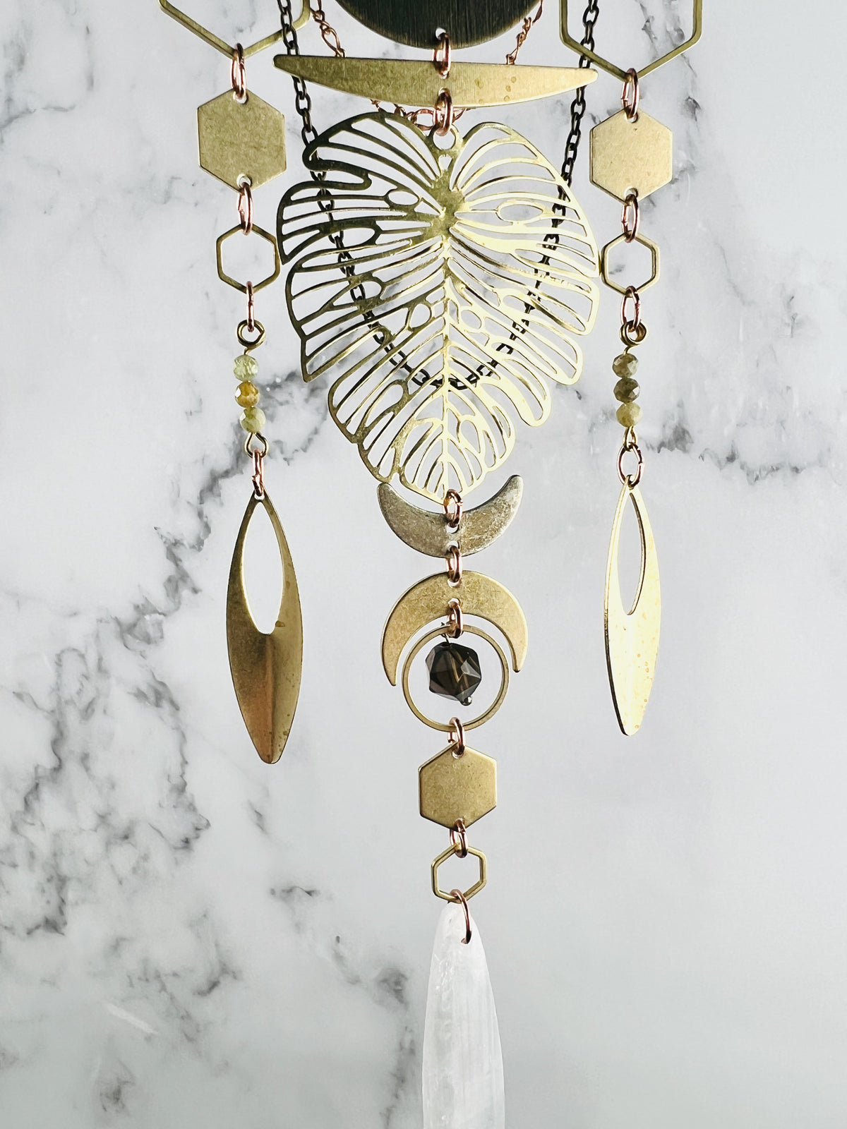 Three sections of vertically hanging, connected geometric shapes and stones adorn this crystal wall art including: hexagons, crescent moons, a large Monstera leaf, ovals, Rings, green garnet and selenite. All hang in front of a gray and white marble wall.