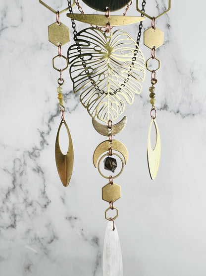 Three sections of vertically hanging, connected geometric shapes and stones adorn this crystal wall art including: hexagons, crescent moons, a large Monstera leaf, ovals, Rings, green garnet and selenite. All hang in front of a gray and white marble wall.