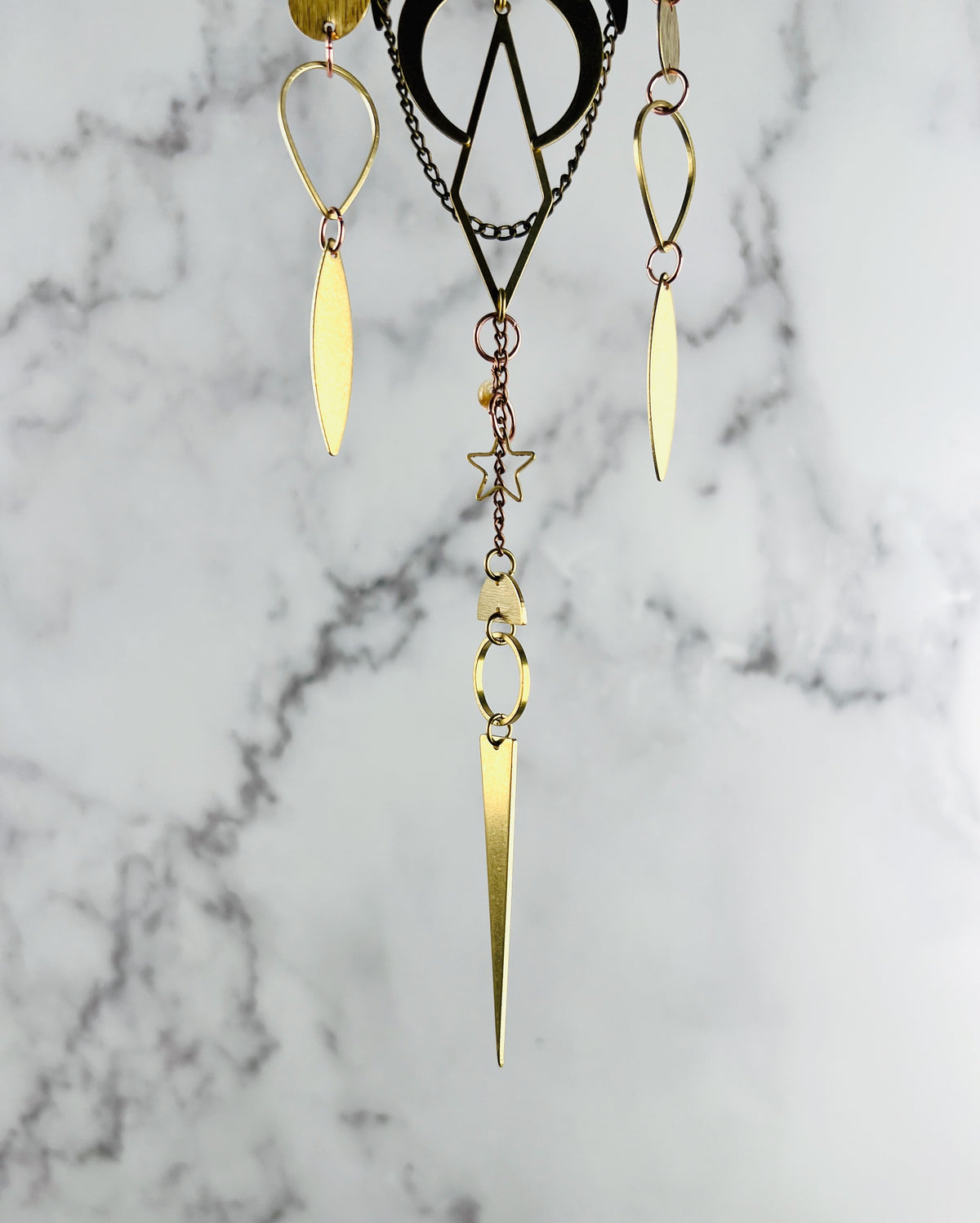 The bottom portion of a piece of crystal wall art depicted as three separate hanging metal patterns and a citrine bead with gold colored  shapes, including: upside down teardrops, diamonds, crescent moon, elongated eye shapes, triangles, half circles, rings, and a star.