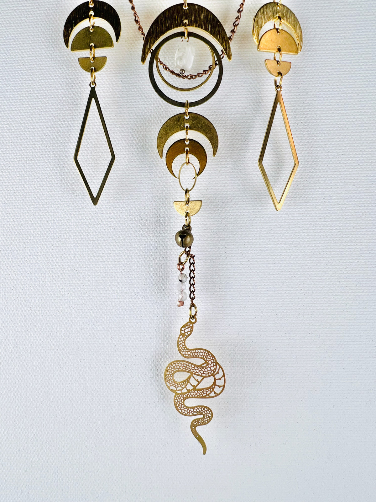 Three distinct sections of vertically hanging, connected gold metal pieces dangle in front of a white canvas wall. Components on it include: a wire wrapped opaque white Bead, copper colored chain, gold colored metal crescent moons, half circles, diamonds, wire wrapped moonstone beads, and rings with a snake Charm and bell at the bottom.