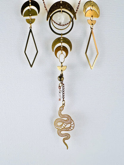 Three distinct sections of vertically hanging, connected gold metal pieces dangle in front of a white canvas wall. Components on it include: a wire wrapped opaque white Bead, copper colored chain, gold colored metal crescent moons, half circles, diamonds, wire wrapped moonstone beads, and rings with a snake Charm and bell at the bottom.