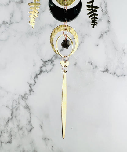 Picture of the bottom of three sections of a vertically hanging piece of dimensional crystal wall art, including: gold colored metal Fern leaves, a black acrylic moon, crescent, moon, rings, half circles and long gold oval at the end, hanging in front of a gray and white marble wall.