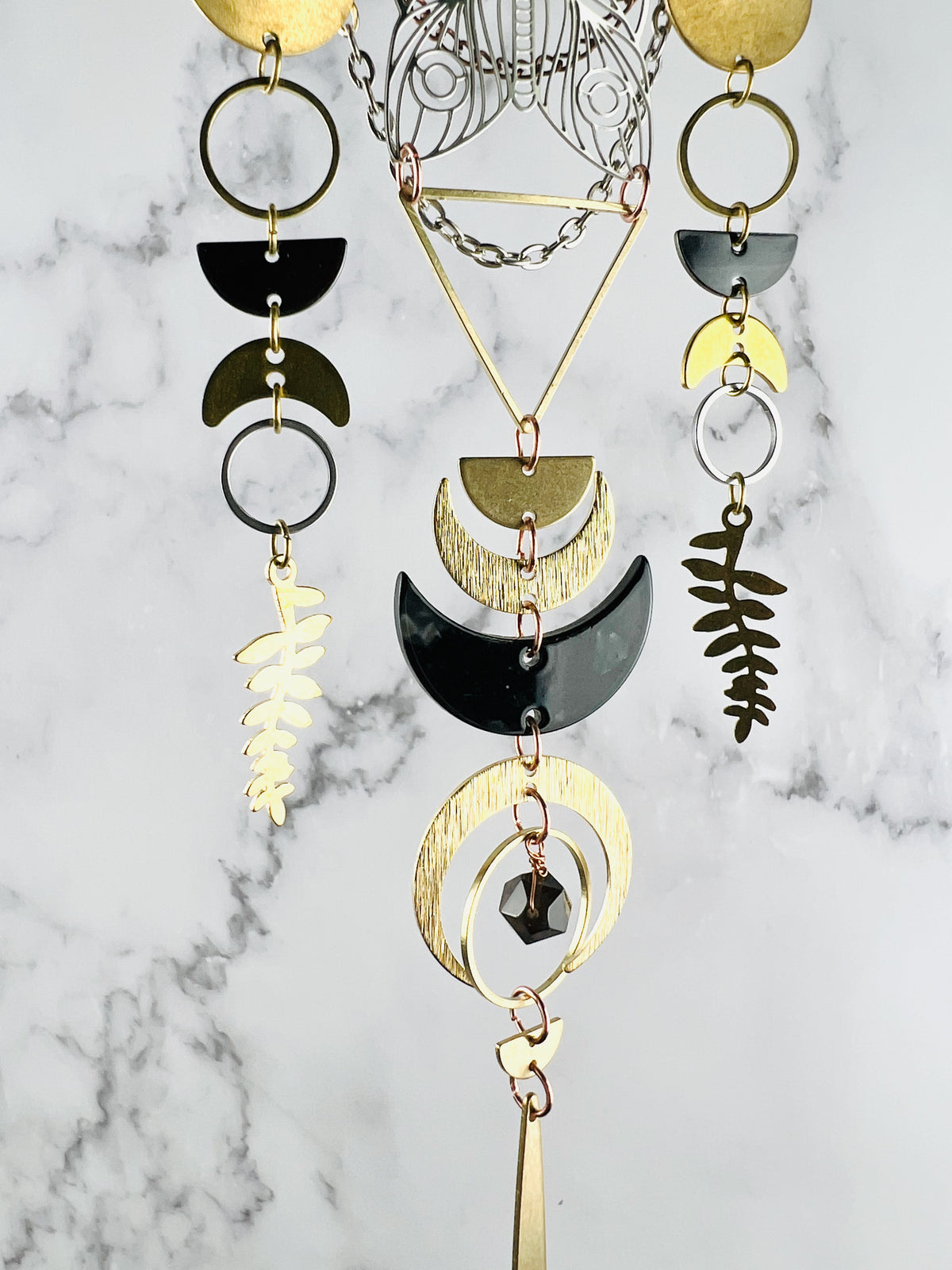 A photo of vertical hanging, three dimensional wall art, depicting wire wrapped crystals metals components, linked with tiny rings. It looks similar to a dream catcher and includes: gold colored, metal, half moons, rings, triangles, crescent moons and a long gold piece at the bottom. There are also silver and bronze colored chains and a stainless steel metal moth cut out. All hang in front of a gray and white marble wall.